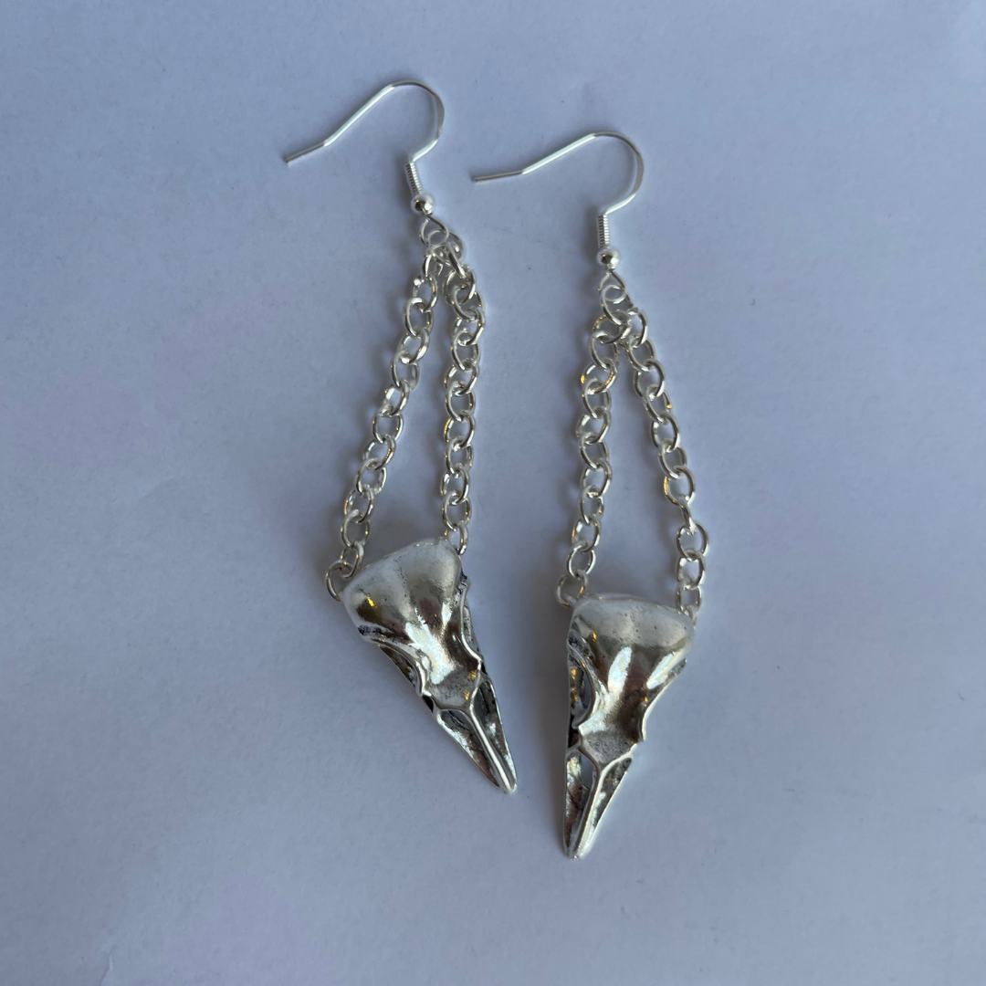 Close-up of handcrafted birdskull charm earrings showing intricate silver chain detail.